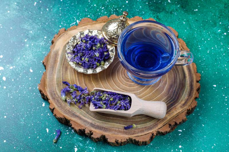 A cup of butterfly pea tea alongside its leaves on a wooden tray