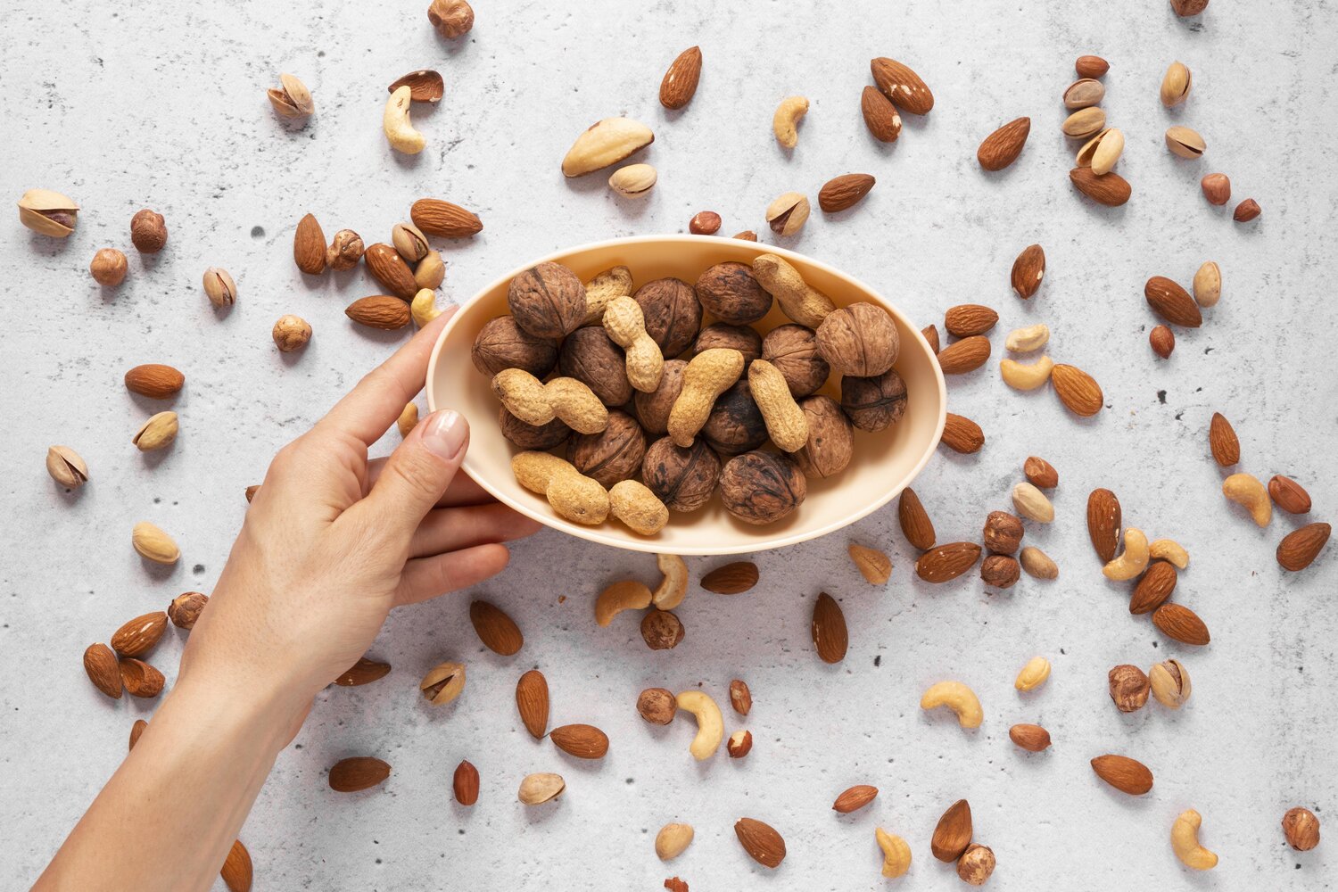 Sebelah tangan memegang mangkuk yang penuh dengan pelbagai jenis kacang untuk diet, dengan lebih banyak kacang tersebar di sekelilingnya.