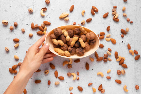 Sebelah tangan memegang mangkuk yang penuh dengan pelbagai jenis kacang untuk diet, dengan lebih banyak kacang tersebar di sekelilingnya.