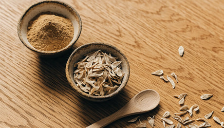 Bowl of ground golpar spice beside whole dried golpar seeds on a wooden table