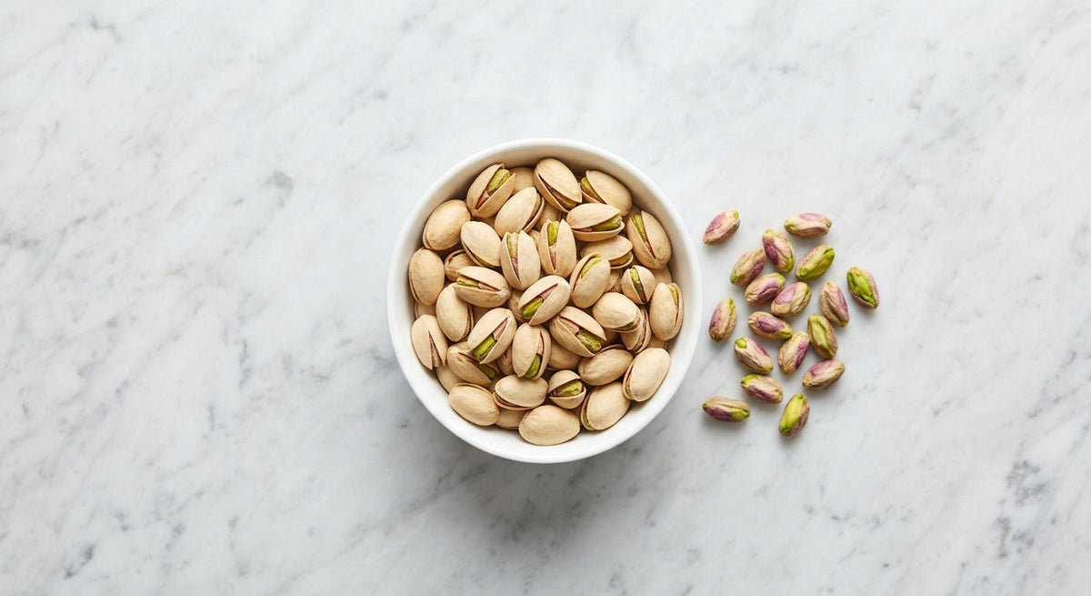Roasted pistachios in their split shells in a white bowl beside loose kernels