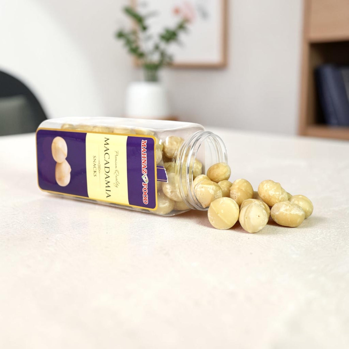Mahnaz Food premium macadamia nuts in a clear plastic jar, spilled out on a white background.