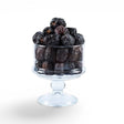 Mahnaz Food Madinah Ajwa dates in a clear glass pedestal bowl, dark brown to black with wrinkles.