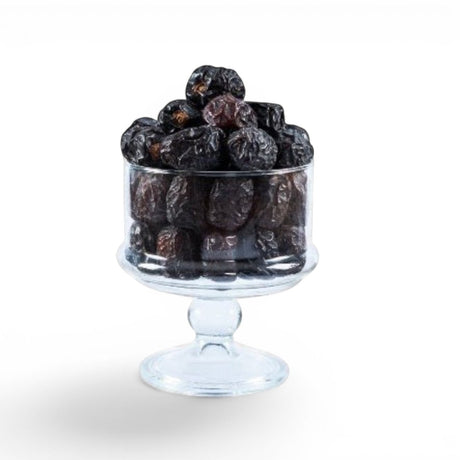 Mahnaz Food Madinah Ajwa dates in a clear glass pedestal bowl, dark brown to black with wrinkles.