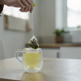 Mahnaz Food Moringa Herbal Tea Bag being steeped in a clear double-walled mug filled with golden tea.