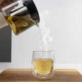 Mahnaz Food Aromatic Natural Nettle Tea being poured into a clear double-walled glass on a wooden cutting board.